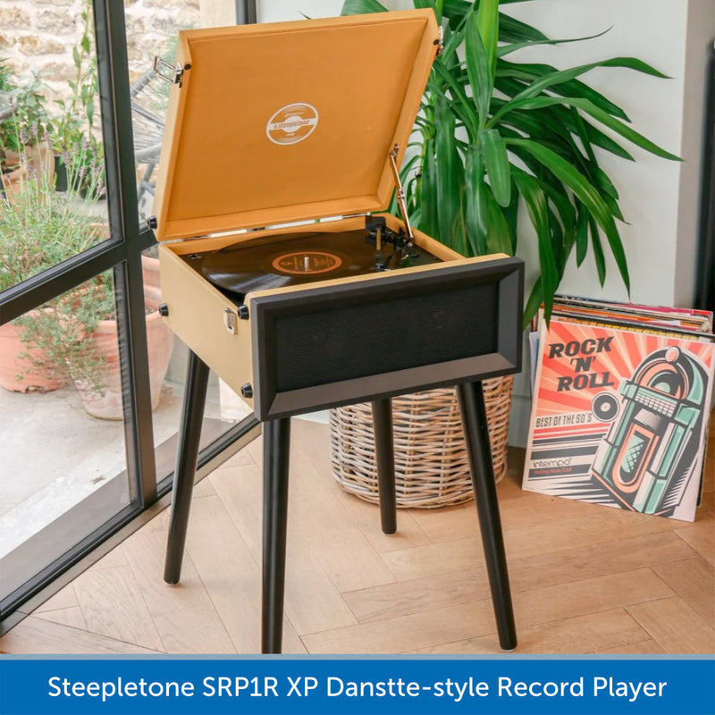 A Steepletona SRP1R XP record player styled in a home environment next to a window, with a plant and vintage-style vinyl records in a basket.